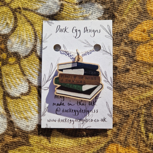 A wooden book stack pendant on a silver chain attached to a white backing card sits on a floral patterned background.