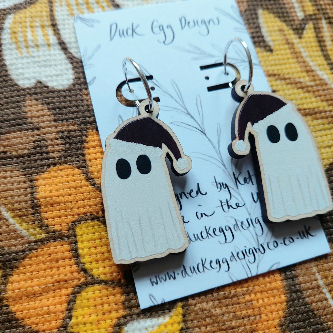 A closer view of the ghost wearing red Christmas hats hang from a pair of silver hoops on a white backing card with the words ‘Duck Egg Designs’ in black handwriting. Behind the backing card you can see brown retro floral patterned fabric.