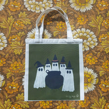Load image into Gallery viewer, A white bag sits on a retro floral patterned background. The bag has a deep green square printed on it with a group of ghosts wearing witches hats stood around a cauldron. 
