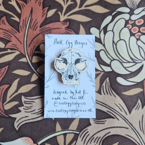 A cat skull wooden pin badge sits on a white and black backing card with the words ‘Duck Egg Designs’ across the top. Behind the backing card you can see a brown retro floral patterned fabric.