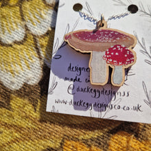 Load image into Gallery viewer, A close up view of the fly agaric necklace showing the detail of the illustrations. The necklace sits on a white backing card with the words ‘Duck Egg Designs’ in black handwriting as well as leafy vines drawn on the card. Behind the backing card you can see a retro floral patterned warm brown fabric.

