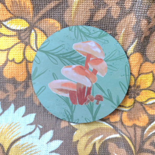 A muted green circle coaster with a velvet shank fungi design sits on a warm brown retro floral patterned fabric. 