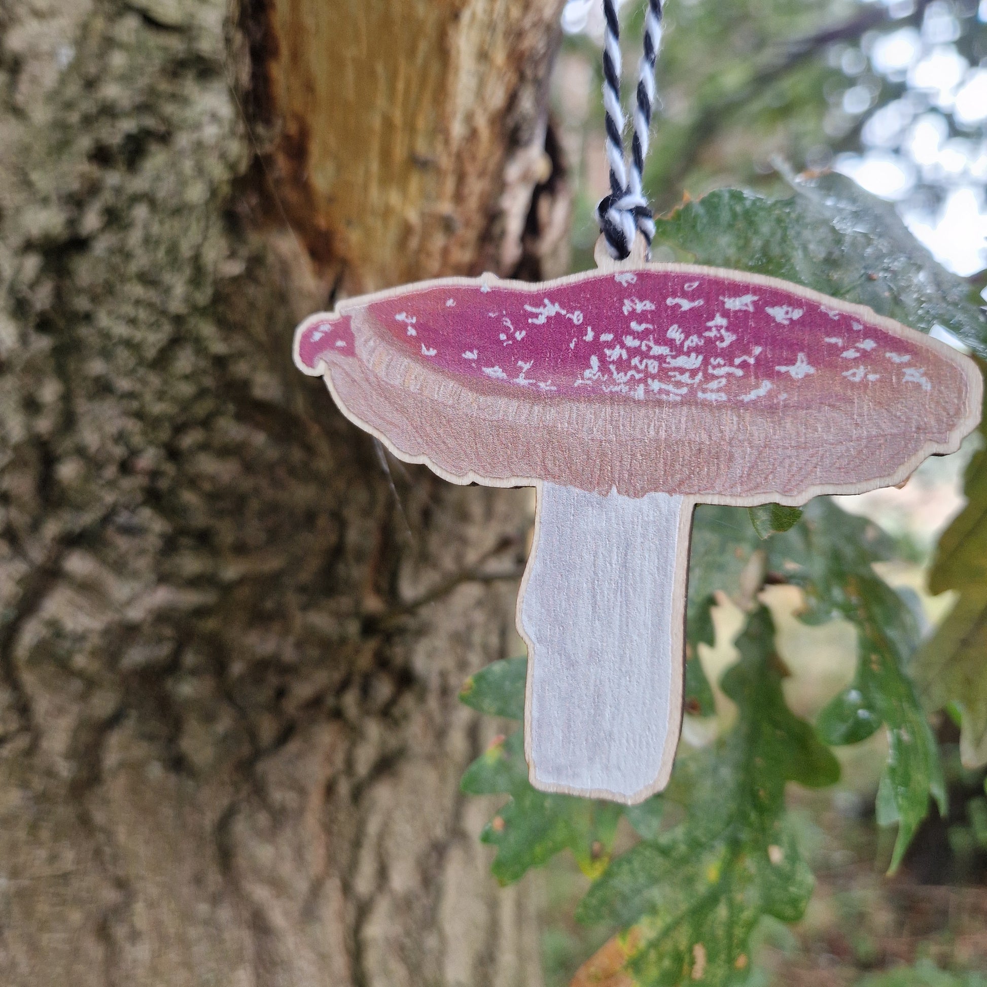 The Fly Agaric Fungi Toadstool Hanging Decoration by Duck Egg Designs, painted as a red-capped mushroom, hangs from a tree branch with a tree trunk and green leaves in the background.