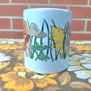 The middle section of the white mug with fungi, grass and autumn leaves on it. The mug sits on a warm retro brown floral fabric and in front of a red brick wall. 