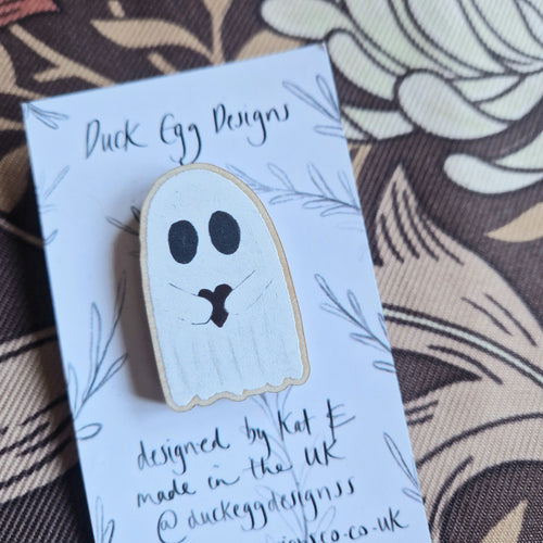 A close up of a wooden ghost pin featuring a ghost holding a red heart. The pin sits on a white and black backing card in front of a brown retro floral background.