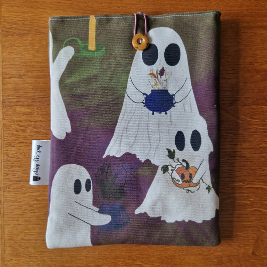 The A Fright Of Ghosts Book Sleeve by Duck Egg Designs features cartoon ghosts performing magic with cauldrons, flowers, and pumpkins on a purple and green background—perfect protection for your favorite books.