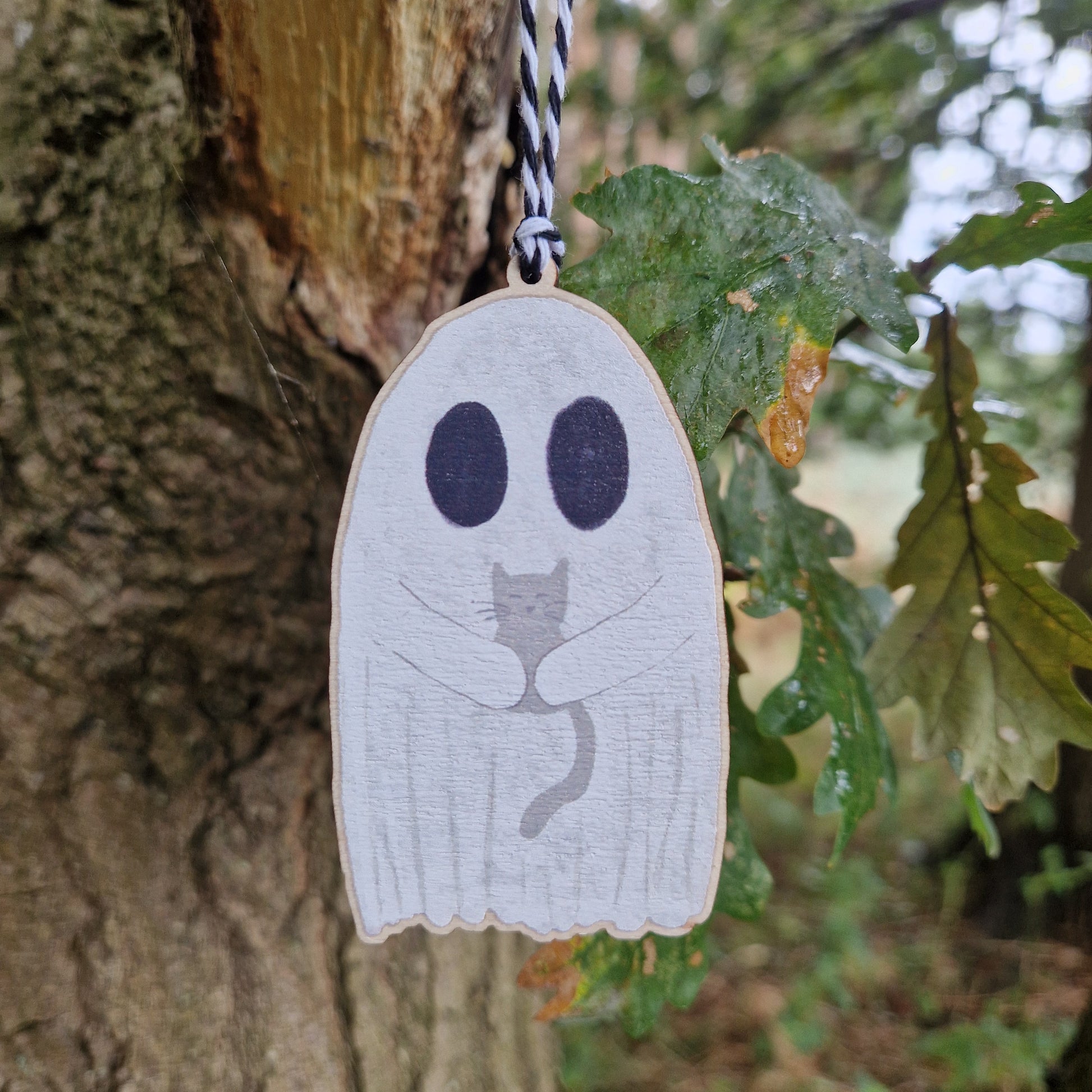 The Ghost With Cat Hanging Wooden Decoration by Duck Egg Designs features a wooden ghost holding a gray cat, hanging from a black-and-white string on a tree branch next to green leaves—perfect for charming wall decor.