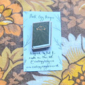 A green pin badge in the shape of a book with the word ‘PLANTS’ in warm yellow writing sits on a white backing card with black writing on it. Behind the backing card is a brown floral patterned background.