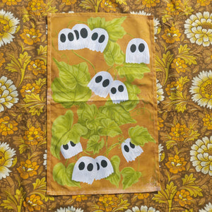 A warm deep orange tea towel with a pumpkin vine design featuring little ghost pumpkins sits on a warm brown retro floral background.