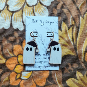  A pair of ghosts wearing dark red Christmas hats hand from a pair of,silver hoops on a white backing card with the words ‘Duck Egg Designs’ in black. In the background you can see a warm brown retro floral patterned fabric.