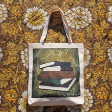Load image into Gallery viewer, A white tote bag sits on a warm brown retro floral patterned background. The tote features a green square with white leafy vines growing inwards from around the edge of the print, as well as a stack of five books which from top to bottom are dark blue, black, brown, green and dark red. 
