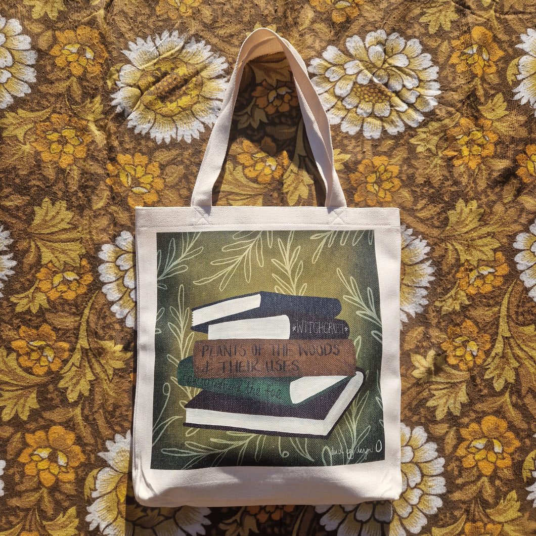 A white tote bag sits on a warm brown retro floral patterned background. The tote features a green square with white leafy vines growing inwards from around the edge of the print, as well as a stack of five books which from top to bottom are dark blue, black, brown, green and dark red. 