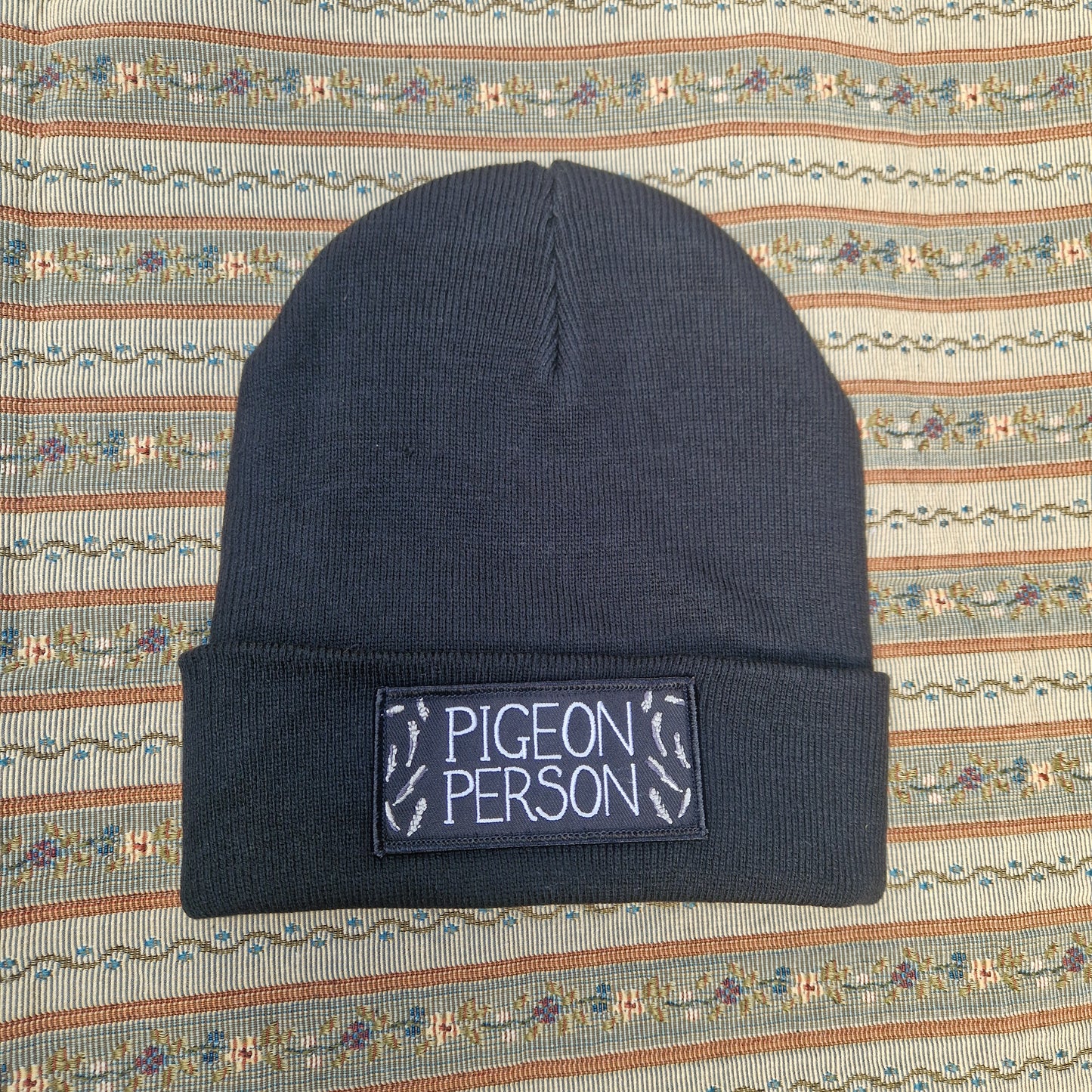 The Pigeon Person Beanie by Duck Egg Designs is a black unisex hat with a folded brim, a patch that reads "PIGEON PERSON," and small pigeon footprints, displayed on patterned fabric.