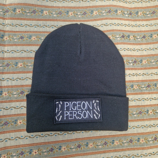 The Pigeon Person Beanie by Duck Egg Designs is a black unisex hat with a folded brim, a patch that reads "PIGEON PERSON," and small pigeon footprints, displayed on patterned fabric.