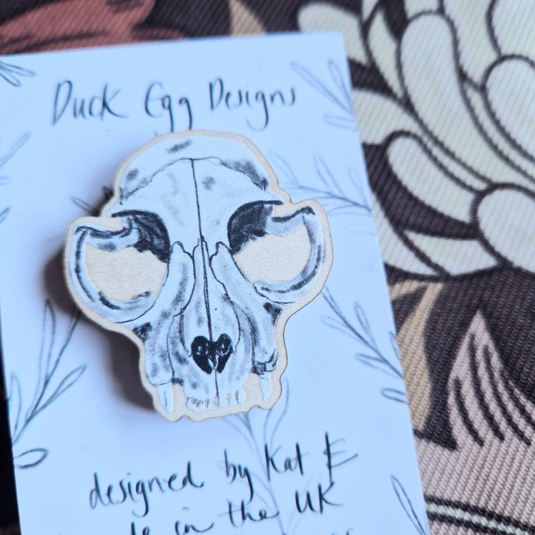 A close up view of a cat skull wooden pin badge sits on a white and black backing card with the words ‘Duck Egg Designs’ across the top. Behind the backing card you can see a brown retro floral patterned fabric.