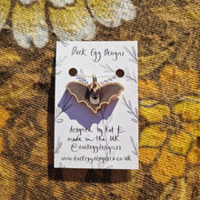 Load image into Gallery viewer, A white backing card sits in the middle of a retro floral brown patterned fabric with a silver chain featuring a brown bat pendant.
