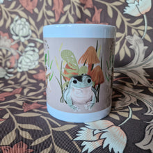 Load image into Gallery viewer, The middle section of a white mug in front of a retro floral patterned background. The mug features a green frog in front of ferns, fungi and grasses Ona peachy coloured background.
