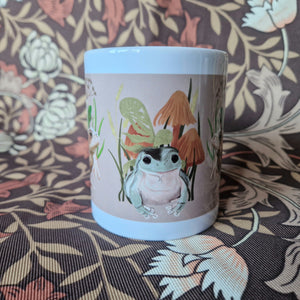 The middle section of a white mug in front of a retro floral patterned background. The mug features a green frog in front of ferns, fungi and grasses Ona peachy coloured background.