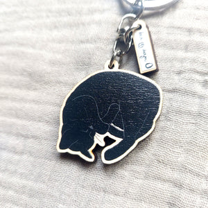 A close up view of the wooden charm on a sleeping black cat key ring. The key ring sits on an off grey fabric background. 