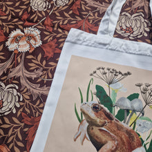 Load image into Gallery viewer, A closer view of the top left of the bag showing the toads upper half as well as fungi, wildflowers and grasses. Above and to the left of the bag you can see a retro brown floral patterned fabric.
