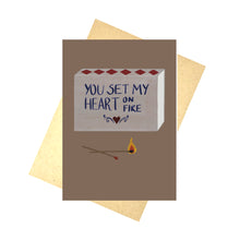 Load image into Gallery viewer, A warm browny purple card with a grey matchbox design on it sits in front of a brown recycled paper envelope on a white background. The match box has words on it that read ‘ You set my heart on fire’ in blue with a red heart and blue symmetrical swirls below it.  In front of the matchbox are two matches, one of which is on fire.
