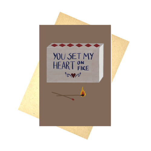 A warm browny purple card with a grey matchbox design on it sits in front of a brown recycled paper envelope on a white background. The match box has words on it that read ‘ You set my heart on fire’ in blue with a red heart and blue symmetrical swirls below it.  In front of the matchbox are two matches, one of which is on fire.