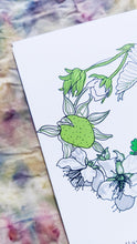 Load image into Gallery viewer, A close up of a corner of the card featuring the green and pink strawberry plant as well as the blossom. Behind the card is naturally dyed fabric featuring warm red, purple, blue and brown tones.
