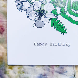 Close up of the bottom left of the card, featuring the words 'Happy Birthday' below the white blossom and green leaves. Behind the card is an abstrac naturally dyed piece of fabric featuring a variety of different tones of red, green, brown, purple and blue.