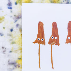 A close up of the card showing the painted texture of the fungi and their simple white and black eyes. Behind the card is a naturally dyed background featuring yellow, blue and purple tones.