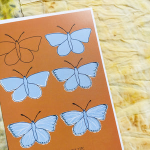 Close up of the Common Blue Butterfly. A warm orange portrait card with six butterflies in rows of two. The butterflies are in the different stages of drawing showing the illustration going from just an outline to the full piece. At the bottom of the card is the english and latin names of the butterfly. Behind the card are naturally dyed fabrics featuring warm yellow, blue and orange tones. - Duck Egg Designs Co