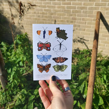 Load image into Gallery viewer, A photo of the card in real life outside, the card is held by a hand with black nail polish and features a sunny garden with a brick fence behind it. The butterflies on the card from L to R are an orange tip, a comma, a peacock, a small white, a common blue, a painted lady, a small copper and a speckled wood.
