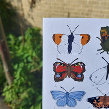 Load image into Gallery viewer, A close up view of the three butterflies on the left of the card ,behind the card a green leafy vine is visible as well as a wooden post and a brick wall. 
