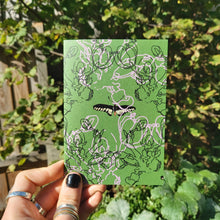 Load image into Gallery viewer, A mossy green card featuring a moth in the middle, with line drawings of blossom overlaying it in light pink and black. The card is being held in the bottom left corner by a hand with black nail polish wearing silver rings. Behind the card a sunny garden with a wooden fence and lots of greenery is visible.
