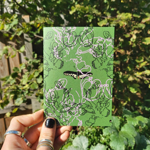 A mossy green card featuring a moth in the middle, with line drawings of blossom overlaying it in light pink and black. The card is being held in the bottom left corner by a hand with black nail polish wearing silver rings. Behind the card a sunny garden with a wooden fence and lots of greenery is visible.