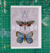 Load image into Gallery viewer, A grey-pink card featuring a layered design created using illustrations from my sketchbook. Central to the card are a rhino beetle, a tropical moth and a blue butterfly. Behind the card is a white envelope, behind which is a green cutting mat with with lines across it.

