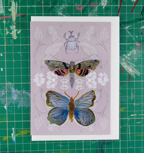 A grey-pink card featuring a layered design created using illustrations from my sketchbook. Central to the card are a rhino beetle, a tropical moth and a blue butterfly. Behind the card is a white envelope, behind which is a green cutting mat with with lines across it.