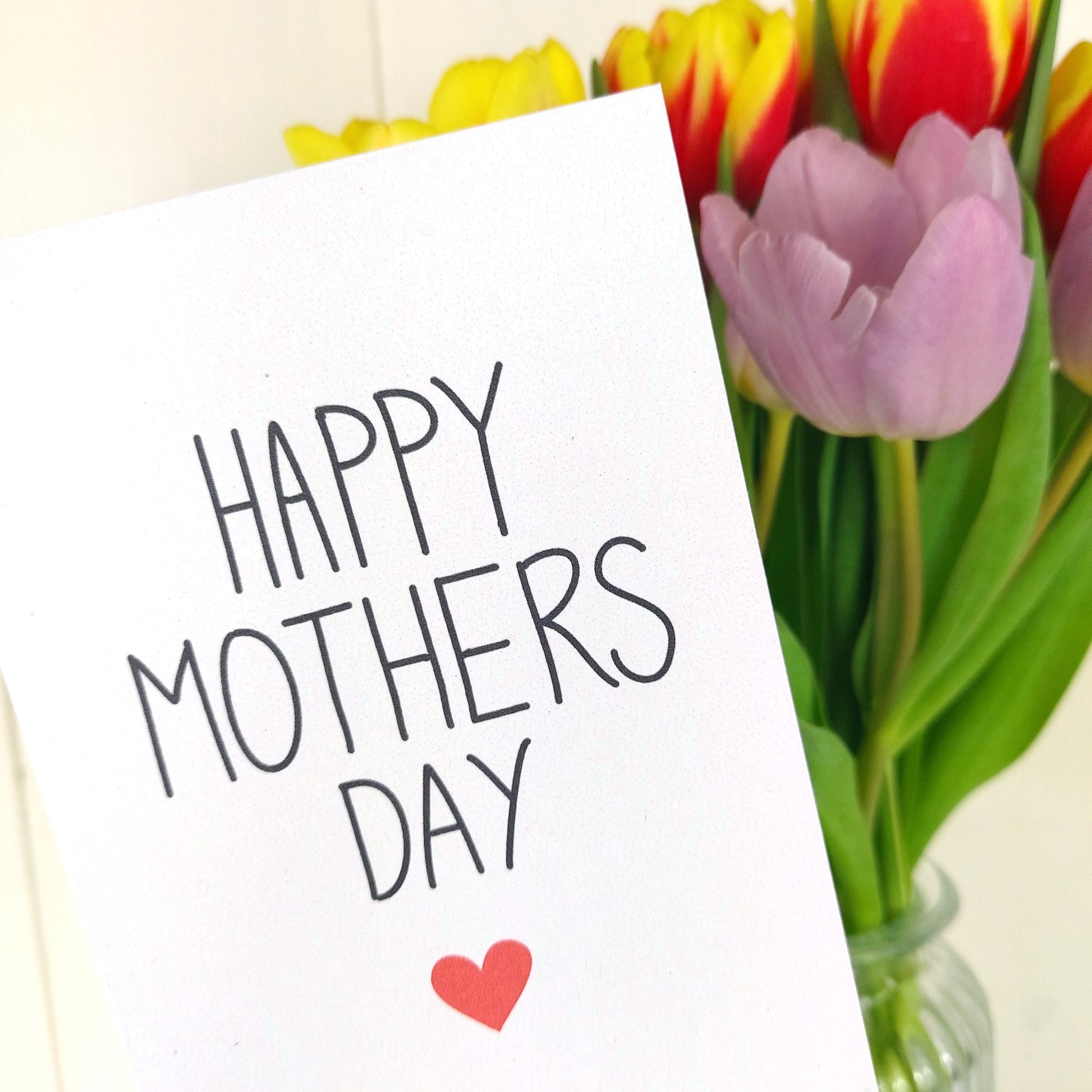 White card at an angle takes up the left side of the photo. The card features the words 'HAPPY MOTHERS DAY' in black writing, below which is a red heart. Behind the card you can see a glass vase of purple and yellow and red tulips. Behind which is a cream wall.