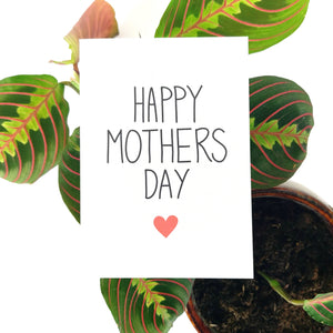 A white card with the words 'HAPPY MOTHERS DAY' in black handdrawn text, central to the card. Underneath the writing is a small red heart, and below the card is a maranta leucona in a brown pot on a white background.