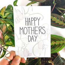 Load image into Gallery viewer, A white card featuring the words HAPPY MOTHERS DAY in black handwriting, with sweet peas behind it in green, pale pink and cream. The card is being held bottom left by a hand with a silver ring on it. Behind the card is an array of houseplants - a hoya carnosa tricolour, a maranta leucona and a ludisia discolor. 
