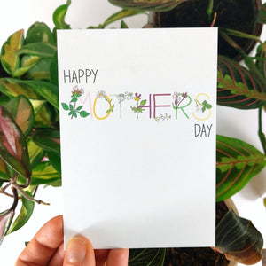 A white card held in front of some colourful houseplants (a hoya carnosa tricolor, a maranta leucona and a ludisia discolor) behind which are a white background. Across the card you can see the words HAPPY MOTHERS DAY the word mother is in colourful writing with the corresponding flowers on the letters.