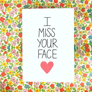 A white card with the words 'I MISS YOUR FACE' down the middle, below which is a red heart. Behind the card is a floral patterned background.