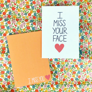 Two cards on a floral background, one on the top right, the other underneath it and towards the bottom left. The right hand card is white with the words 'I MISS YOUR FACE' in black with a red heart underneath. The card on the left is a warm orange with the words 'I MISS YOU' in white next to a red heart.