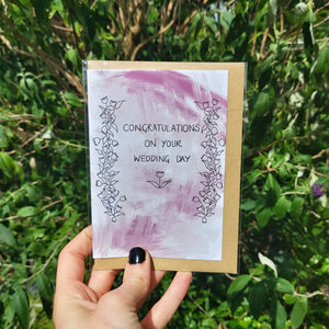 Simple wedding card with a textured pastel red/pink background. Black floral vines line the left and right of the card, with a flower with leaves visible below the words 'Congratulations On Your Wedding Day'. The card is in a compostable sleeve with a recycled brown envelope. The card is held by a hand with black nail polish in front of a leafy green plant.