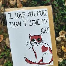 Load image into Gallery viewer, A close up view of the card, showing a ginger and white cat with a love heart marking on its side, and green eyes. Above the cat are the words &#39;I LOVE YOU MORE THAN I LOVE MY CAT&#39; in black writing. The card is in a compostable sleeve and you can see a recycled brown envelope in with it. Behind the card you can see a sunny overgrown garden.
