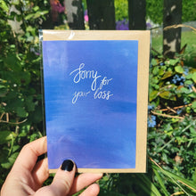 Load image into Gallery viewer, A hand with black nail polish holds a blue card with the words &#39;Sorry for your loss&#39; in white calligraphy. The card is in a compostable sleeve, with a brown recycled envelope visible behind it. Behind the card is an overgrown green sunny garden.
