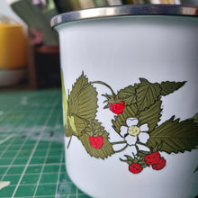 Load image into Gallery viewer, Close up of the white enamel cup, showing the deep green wild strawberry plant illustration. The cup is sat on a green cutting mat in front of some books.
