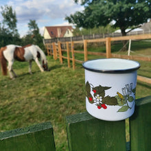 Load image into Gallery viewer, Picture showing the enamel cup in the foreground to the right. The cup is white with a metal rim, and features a print of wild strawberries and honeysuckle. Below the mug is a green wooden fence, behind which you can see a grassy paddock with a chestnut and white horse as well as a tree.
