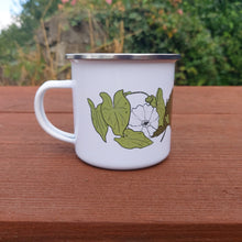 Load image into Gallery viewer, View of the white enamel cups with the handle on the left. You can see the bindweed illustration and part of the strawberry plant illustration. The mug is sat on a wooden picnic bench in front of an overgrown wall.
