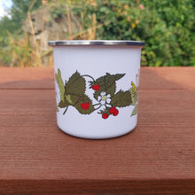 Load image into Gallery viewer, View of the mug with the handle at the back (out of view). You can see the wild strawberry plant illustration, and parts of the other plants. Under the mug is a brown wooden picnic bench, behind which is an overgrown wall.
