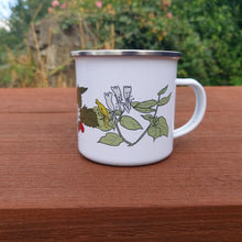 Load image into Gallery viewer, The white mug is shown with the handle on the right, showing the honeysuckle illustration on the cup as well as part of the strawberry plant. The mug is sat on a brown wooden table, behind which you can see an overgrown wall.

